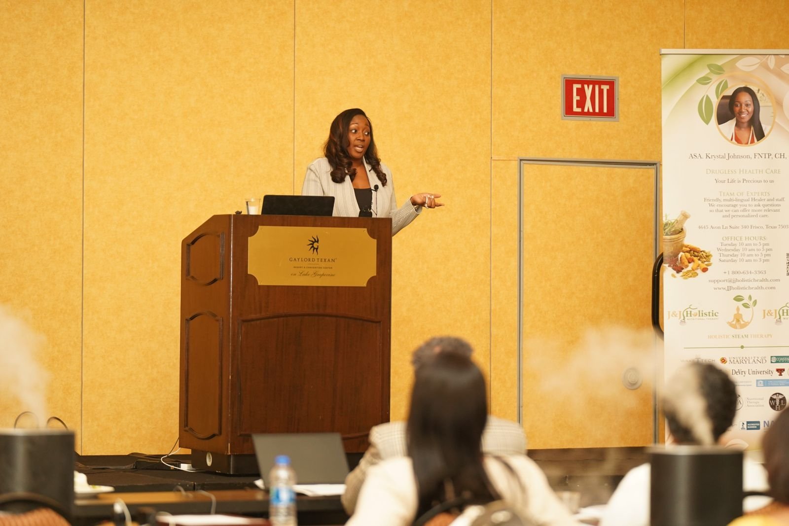 Krystal Johnson delivering a keynote at the Gaylord Texan Resort and Convention Center, speaking to a full corporate audience in her white blazer with J&J Holistic banners in the background
